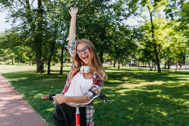 Graziosa ragazza bionda che esprime eccitazione. Foto all'aperto di felice signora bianca con la bicicletta in posa sul parco.