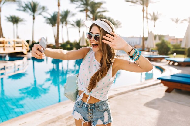 Graziosa ragazza che balla in braccialetti alla moda e cappello bianco che fa selfie prima di nuotare nella piscina all'aperto.