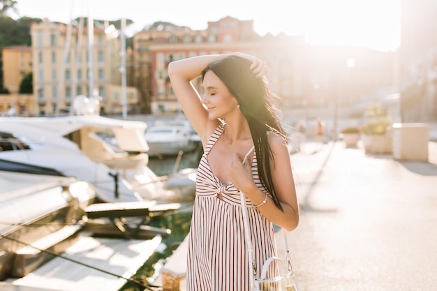 Graziosa ragazza con capelli neri dritti che si rilassano all'aperto e si godono il sole con gli occhi chiusi. Ritratto di elegante signora bruna in posa con splendidi edifici sullo sfondo durante il viaggio.