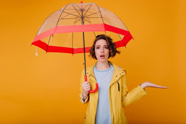 Graziosa ragazza indossa elegante cappotto autunnale in piedi sotto l'ombrellone. Ritratto dello studio del modello femminile caucasico sconvolto che posa con l'ombrello sulla parete gialla.