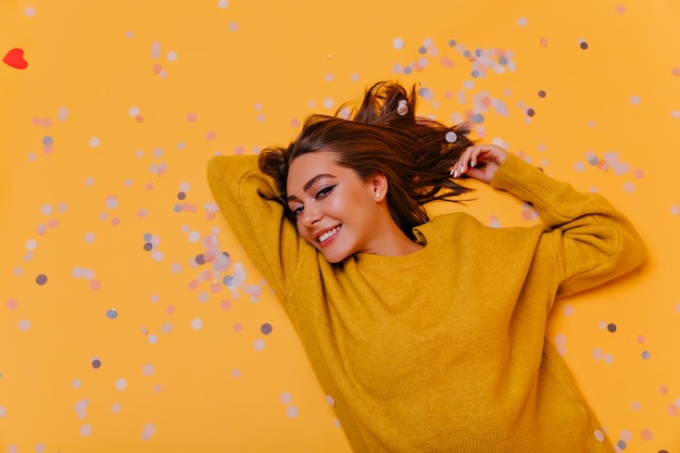 Graziosa signora con capelli castani sdraiato sul muro giallo. Ritratto ambientale della donna alla moda sensuale in maglione arancione.