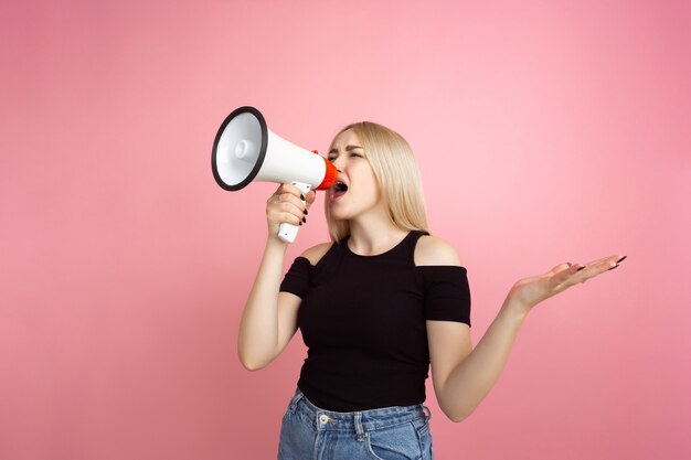 Gridando con il megafono. Ritratto di giovane donna con emozioni luminose sulla parete dello studio rosa corallo