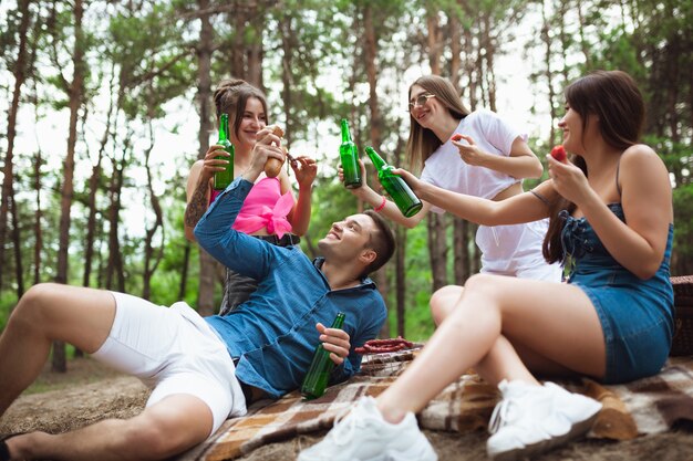 Gruppo di amici che tintinnano bottiglie di birra durante il picnic nell'amicizia dello stile di vita della foresta estiva summer