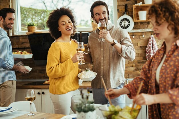 Gruppo di amici felici che preparano cibo e bevono vino in cucina. Il focus è sul giovane con il bicchiere di vino.