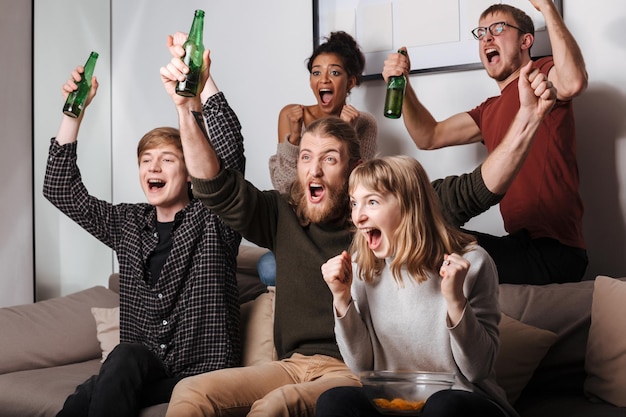 Gruppo di amici gioiosi seduti sul divano e urlando allegramente mentre guardano la tv insieme a patatine e birra a casa