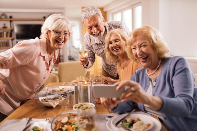 Gruppo di anziani felici che usano lo smartphone e si fanno selfie al tavolo da pranzo a casa