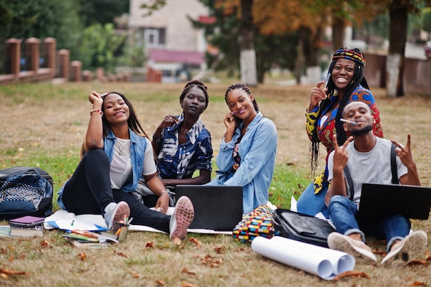 Gruppo di cinque studenti universitari africani che trascorrono del tempo insieme nel campus nel cortile dell'università Amici afro neri seduti sull'erba e che studiano con i laptop