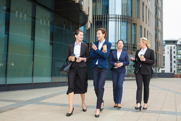 Gruppo di colleghe che camminano con caffè da asporto all'aperto, parlando, sorridendo. A figura intera, vista frontale. Concetto di pausa di lavoro