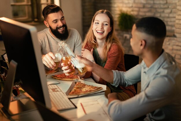 Gruppo di colleghi creativi felici che si divertono mentre brindano con birra e mangiano pizza in ufficio