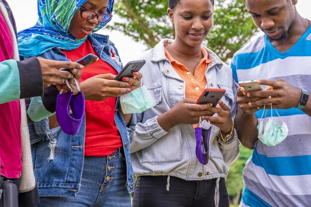 Gruppo di giovani amici africani con maschere che usano i loro telefoni in un parco