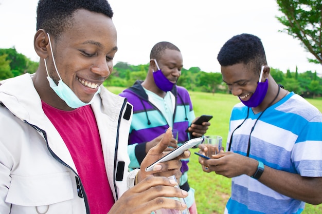 Gruppo di giovani amici africani con mascherine che usano i telefoni mentre si allontanano socialmente in un parco