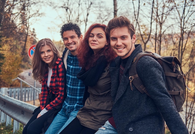 Gruppo di giovani amici con zaini seduti sul guardrail vicino alla strada con una bellissima foresta e un fiume sullo sfondo. Viaggio, escursione, concetto.