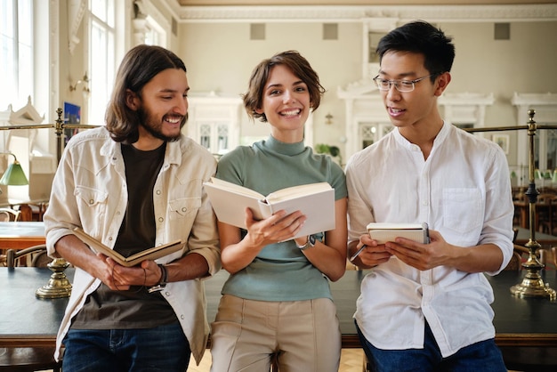 Gruppo di giovani studenti multinazionali casuali che studiano con gioia con un libro e prendono appunti insieme nella biblioteca dell'università