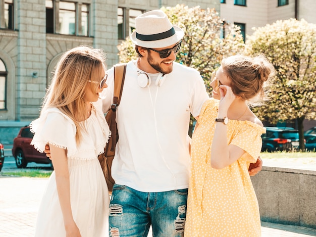 Gruppo di giovani tre amici alla moda che posano nella via. Moda uomo e due ragazze carine vestite in abiti estivi casual. Modelle sorridenti divertendosi in occhiali da sole. Donne allegre e ragazzo che chiacchierano