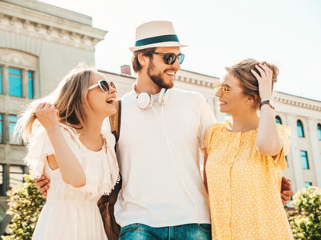 Gruppo di giovani tre amici alla moda che posano nella via. Moda uomo e due ragazze carine vestite in abiti estivi casual. Modelle sorridenti divertendosi in occhiali da sole. Donne allegre e ragazzo che chiacchierano