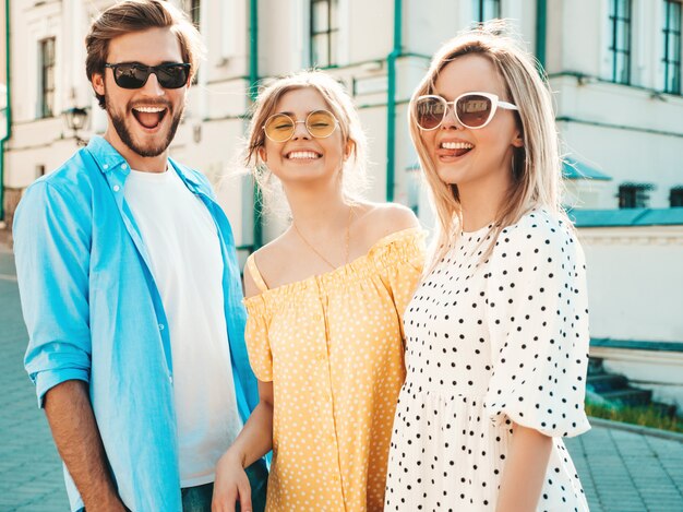Gruppo di giovani tre amici alla moda che posano nella via. Moda uomo e due ragazze carine vestite in abiti estivi casual. Modelli sorridenti divertendosi in occhiali da sole. Donne allegre e ragazzo a susnet