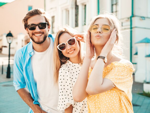 Gruppo di giovani tre amici alla moda che posano nella via. Moda uomo e due ragazze carine vestite in abiti estivi casual. Modelli sorridenti divertendosi in occhiali da sole. Donne allegre e ragazzo a susnet