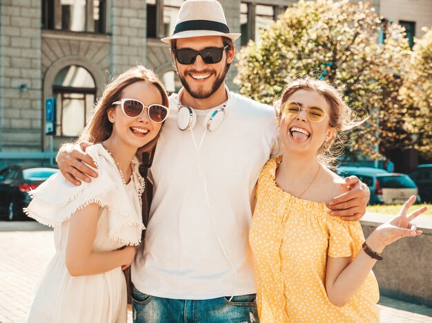 Gruppo di giovani tre amici alla moda che posano nella via. Moda uomo e due ragazze carine vestite in abiti estivi casual. Modelli sorridenti divertendosi in occhiali da sole. Donne allegre e ragazzo che impazziscono
