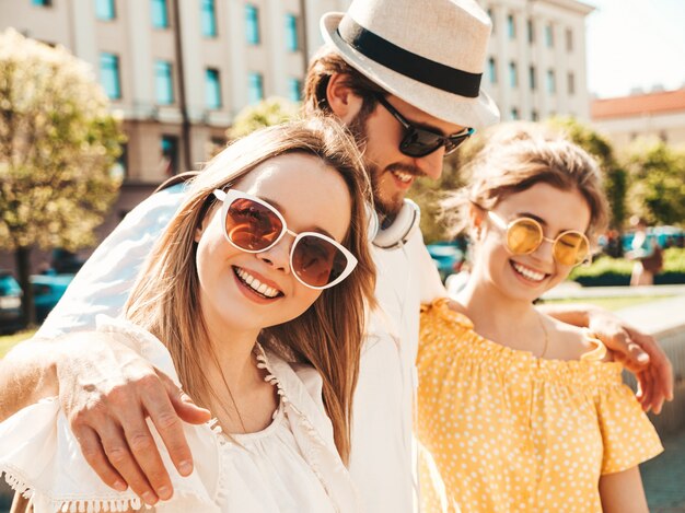 Gruppo di giovani tre amici alla moda che posano nella via. Moda uomo e due ragazze carine vestite in abiti estivi casual. Modelli sorridenti divertendosi in occhiali da sole. Donne allegre e ragazzo che impazziscono