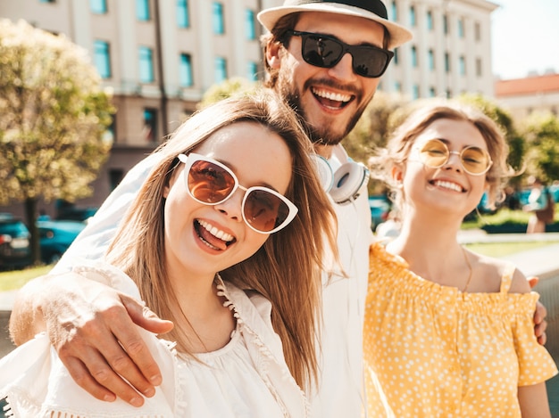 Gruppo di giovani tre amici alla moda che posano nella via. Moda uomo e due ragazze carine vestite in abiti estivi casual. Modelli sorridenti divertendosi in occhiali da sole. Donne allegre e ragazzo che impazziscono