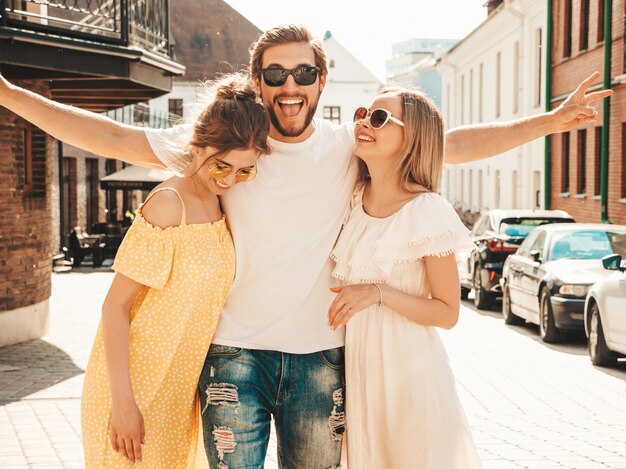 Gruppo di giovani tre amici alla moda che posano nella via. Moda uomo e due ragazze carine vestite in abiti estivi casual. Modelli sorridenti divertendosi in occhiali da sole. Donne allegre e ragazzo che impazziscono