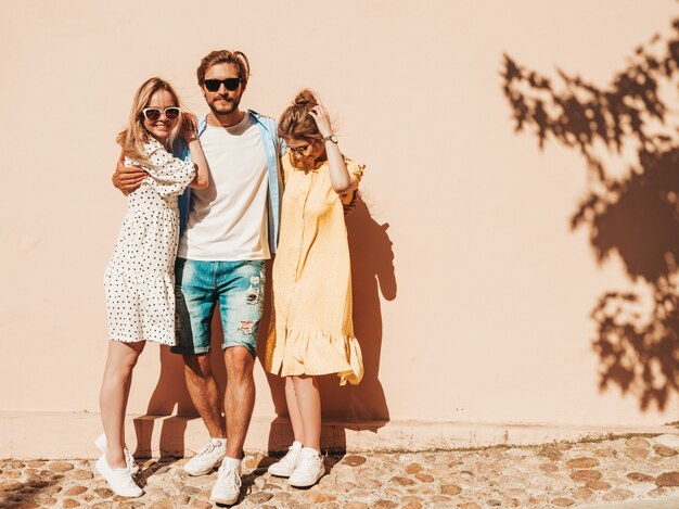 Gruppo di giovani tre amici alla moda che posano nella via. Moda uomo e due ragazze carine vestite in abiti estivi casual. Modelli sorridenti divertendosi in occhiali da sole. Donne allegre e ragazzo che impazziscono