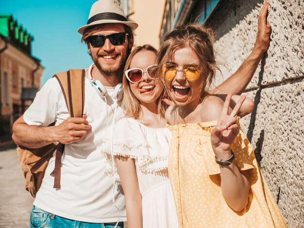 Gruppo di giovani tre amici alla moda che posano nella via. Moda uomo e due ragazze carine vestite in abiti estivi casual. Modelli sorridenti divertendosi in occhiali da sole. Donne allegre e tipo all'aperto