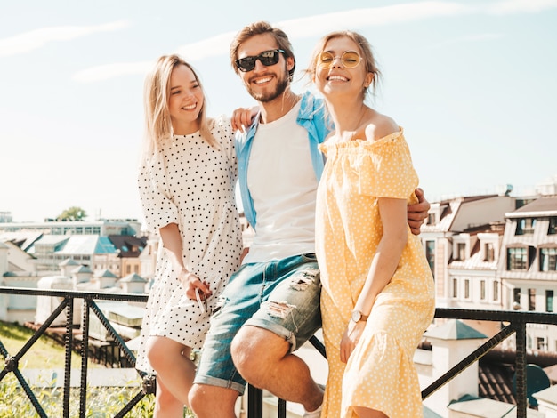 Gruppo di giovani tre amici alla moda che posano nella via. Moda uomo e due ragazze carine vestite in abiti estivi casual. Modelli sorridenti divertendosi in occhiali da sole. Donne allegre e tipo all'aperto