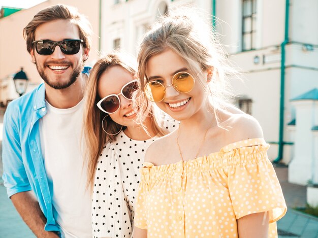 Gruppo di giovani tre amici alla moda che posano nella via. Moda uomo e due ragazze carine vestite in abiti estivi casual. Modelli sorridenti divertendosi in occhiali da sole. Donne allegre e tipo all'aperto