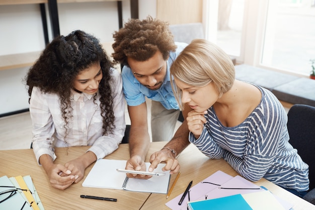 Gruppo di tre giovani imprenditori che lavorano insieme per un nuovo progetto di avvio. Giovani seduti in biblioteca guardando attraverso le informazioni sullo smartphone.