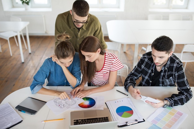 Gruppo di uomini e donne premurosi che lavorano con schizzi e laptop mentre trascorrono del tempo al lavoro Giovani creativi che discutono insieme di nuovi progetti in un ufficio moderno