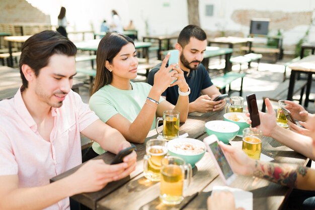 Gruppo eterogeneo di amici sui 20 anni seduti insieme in un bar all'aperto mentre controllano i social media e inviano messaggi sui loro smartphone