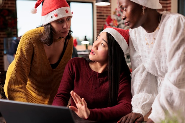 Gruppo eterogeneo di donne che lavorano alla pianificazione aziendale in ufficio festivo con albero di natale e decorazioni. Fare lavoro di squadra e festeggiare il periodo natalizio con addobbi natalizi stagionali.