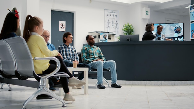 Gruppo eterogeneo di persone in attesa nella hall della reception dell'ospedale per partecipare all'appuntamento medico con il medico generico. Pazienti nella hall della sala d'attesa seduti alla clinica sanitaria. Scatto da treppiede.