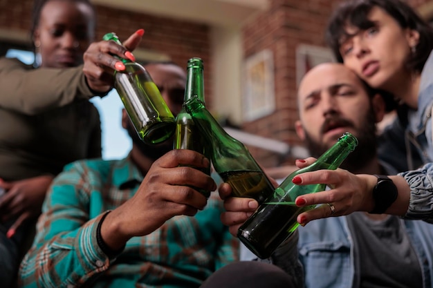 Gruppo multietnico di amici che saluta e tintinnano bottiglie di birra per celebrare l'evento della festa, felice della riunione. Persone che brindano con bevande alcoliche al ritrovo, attività ricreative. Avvicinamento.