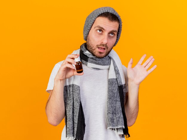 Guardando a lato il giovane malato che indossa cappello invernale e sciarpa tenendo la medicina in una bottiglia di vetro e diffondere la mano isolata su sfondo giallo