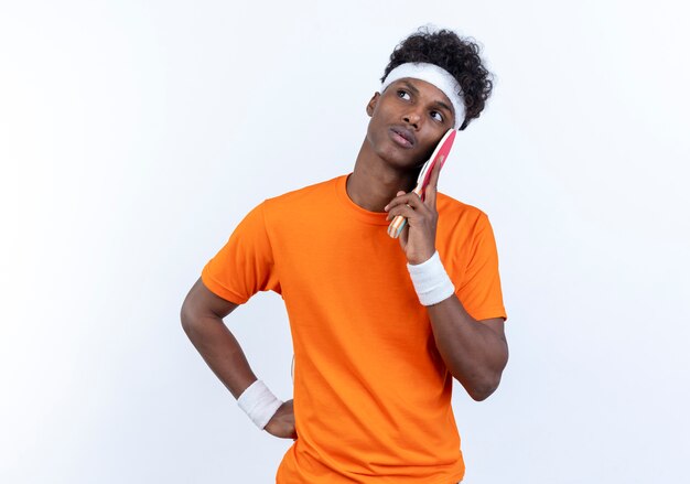 Guardando a lato pensando giovane afro-americano sportivo uomo che indossa la fascia e il braccialetto mettendo la racchetta da ping pong sulla guancia isolati su sfondo bianco