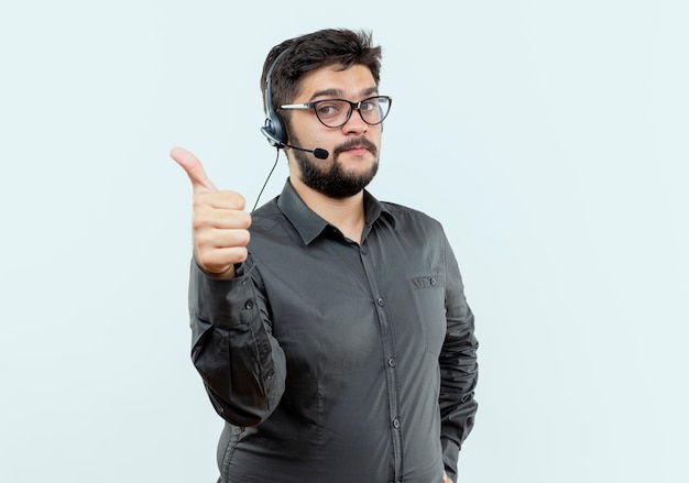 Guardando a porte chiuse giovane call center uomo che indossa auricolare e occhiali il pollice in alto isolato su sfondo bianco