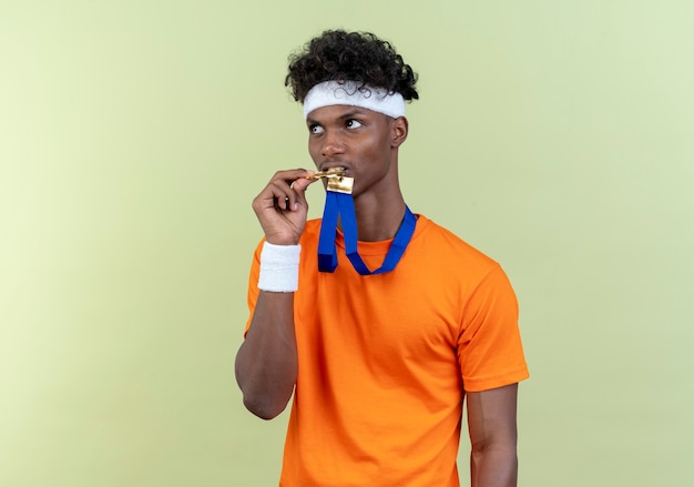 Guardando al lato giovane afro-americano sportivo uomo che indossa la fascia e la fascia da polso holding e morde la medaglia sul collo isolato su sfondo verde