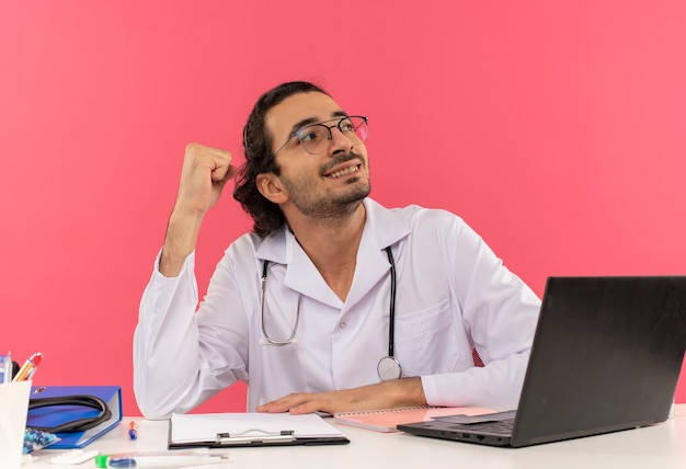 Guardando al lato sorridente giovane medico maschio con occhiali medici che indossano accappatoio medico con stetoscopio seduto alla scrivania