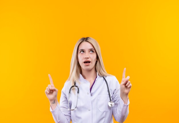 Guardando al medico giovane ragazza che indossa uno stetoscopio in abito medico punta su sfondo giallo isolato