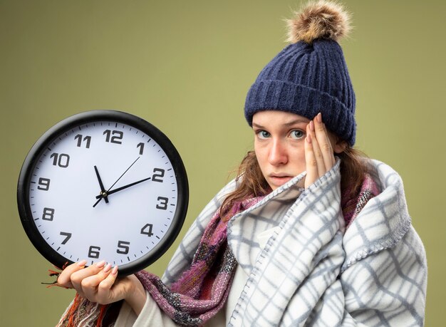 Guardando dritto davanti a sé giovane ragazza malata che indossa una veste bianca e cappello invernale con sciarpa tenendo l'orologio da parete avvolto in un plaid mettendo la mano sul tempio isolato su verde oliva