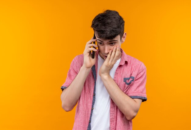 Guardando giù il giovane caucasico che indossa la camicia rosa parla al telefono mise la mano sulla guancia sul muro arancione isolato