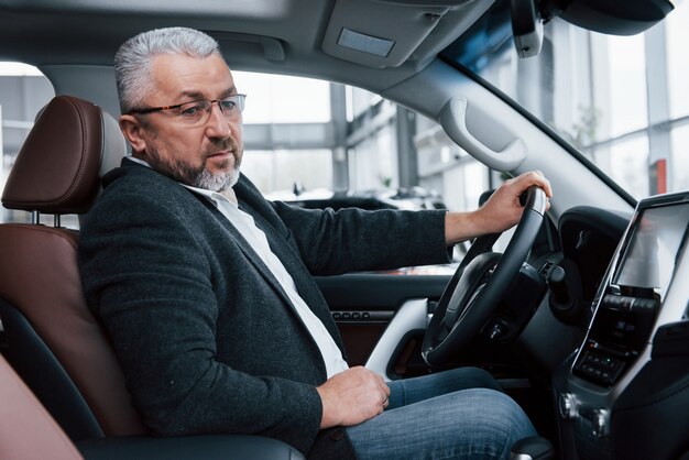 Guardando il design. Uomo d'affari senior in vestiti ufficiali che provano nuova automobile di lusso in salone automatico