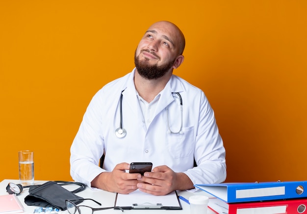 Guardando il felice giovane maschio calvo medico indossando abito medico e stetoscopio seduto alla scrivania con strumenti medici tenendo il telefono isolato su sfondo arancione