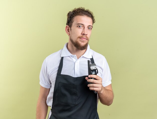 Guardando il giovane barbiere maschio che indossa l'uniforme che tiene le tosatrici con pettine isolato su parete verde oliva