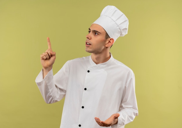 Guardando il giovane cuoco maschio che indossa l'uniforme del cuoco unico punta il dito verso l'alto sulla parete verde isolata con lo spazio della copia