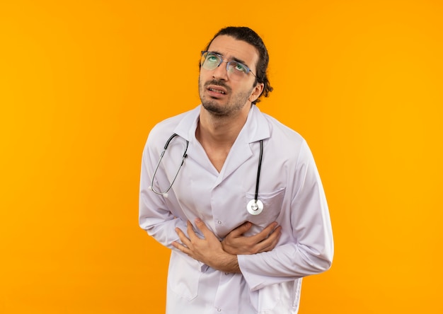 Guardando il giovane medico malato con gli occhiali medici che indossa la veste medica con lo stetoscopio ha afferrato lo stomaco