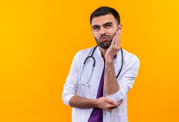 Guardando il giovane medico maschio che indossa l'abito medico dello stetoscopio mise la mano sulla guancia su sfondo giallo isolato