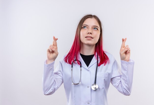 Guardando il giovane medico ragazza che indossa stetoscopio abito medico dita incrociate su sfondo bianco isolato